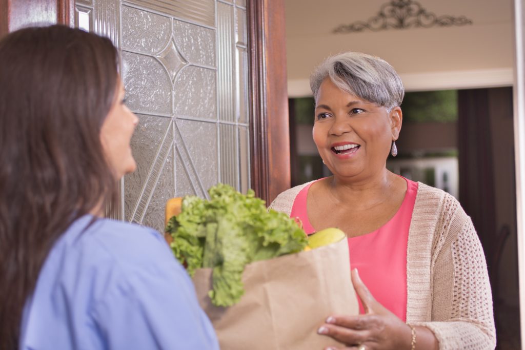 Nurse or female volunteer delivers groceries to a senior adult woman at her home.  The Latin descent woman carries a grocery bag full of fresh fruits and vegetables to the mature woman who is unable to leave her home due to illness.  The woman is grateful for the delivery.
